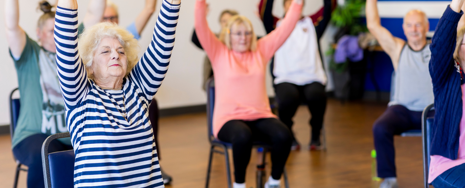 Group of seniors sitting on chairs reaching their arms up to the air. Women in foreground is wearing a blue and white striped top.